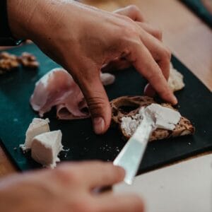 A person spreads soft cheese onto a slice of bread with a knife, next to pieces of gourmet food like cheese, ham, and walnuts on a black cutting board.