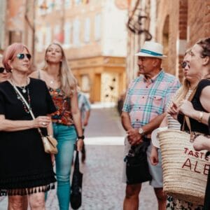 A group of casually dressed people stand and talk on a cobblestone street in a sunny, historic town during a Taste of Albi food tour. Some wear sunglasses and hats; one holds a straw bag. Brick buildings and warm lighting create a relaxed atmosphere.