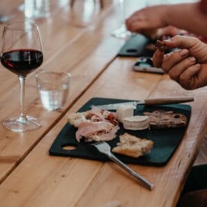 A wooden table with a black cutting board holding cheese, ham, bread, and a fork. A glass of red wine hints at the flavors of an Albi Gourmet Food Tour, while hands prepare to eat beside a refreshing glass of water.