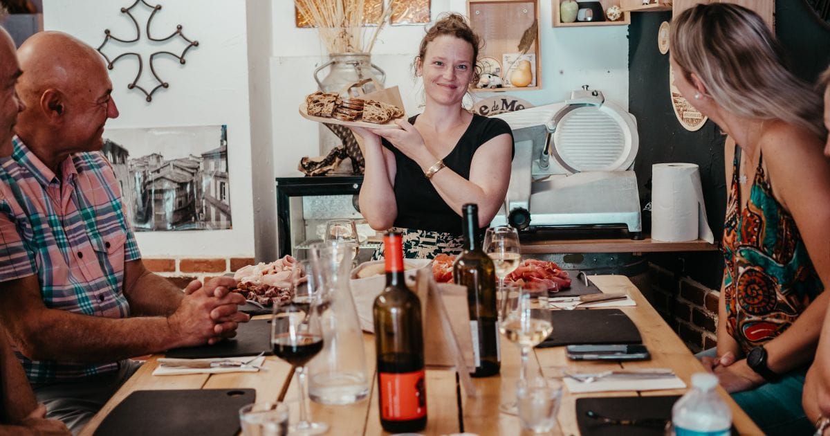 A woman smiles and holds up a plate of food while seated at a wooden dining table with three others during an Albi Gourmet Food Tour. The cozy, rustic setting features wine, glasses, and assorted dishes that capture the region’s authentic flavors.