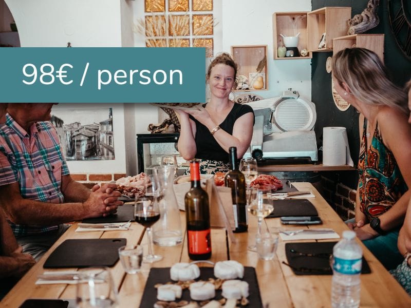 Dans un restaurant toulousain confortable, une femme sourit devant un plateau de nourriture, entourée de trois personnes assises en train de déguster des dégustations. Du vin, de l'eau et du fromage sont posés sur la table. Le texte superposé indique "98€ / personne".