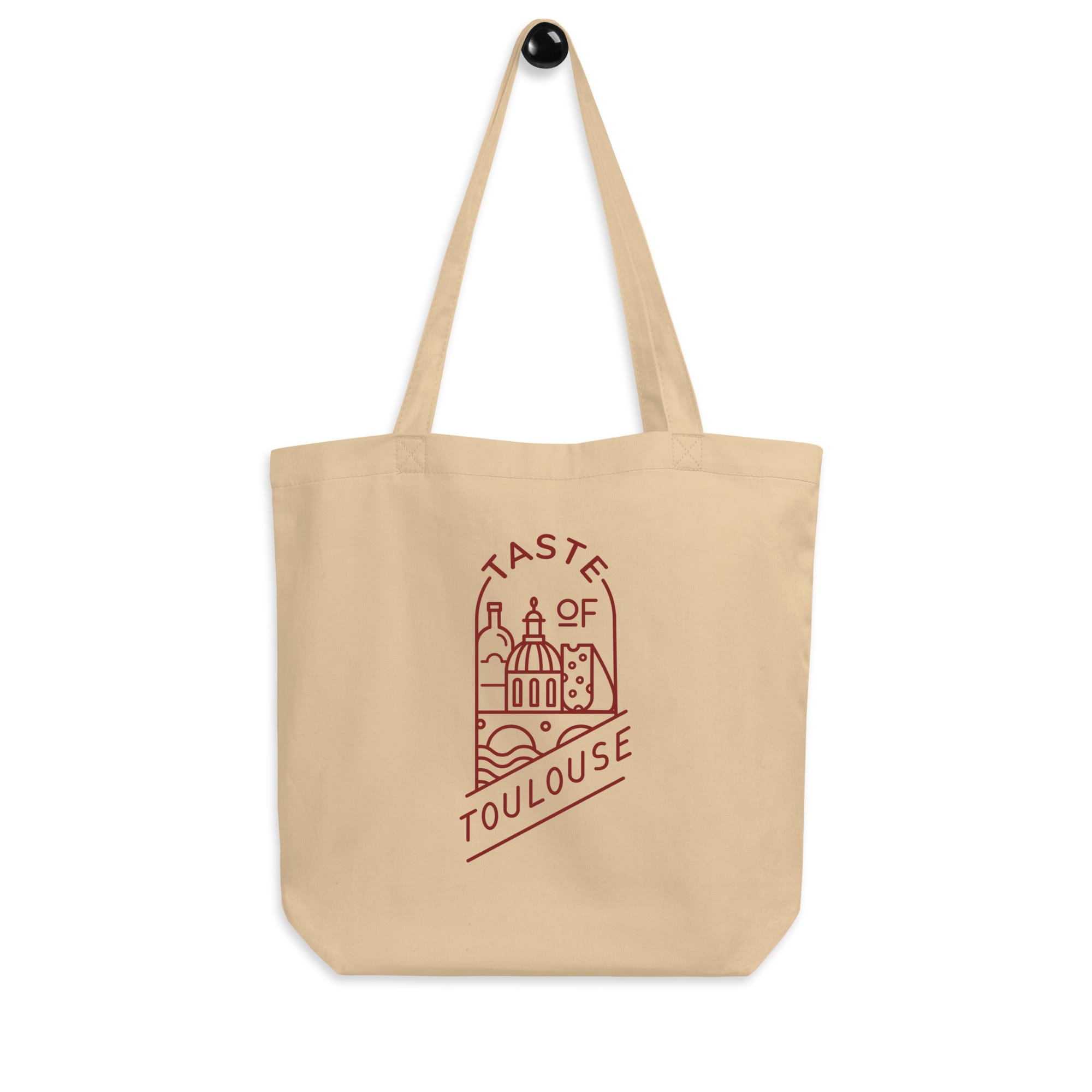 The Tote Bag with Red Logo is a beige tote hanging on a wall, displaying a minimalist red logo and line drawing of Toulouse landmarks, plus "Taste of Toulouse" text.