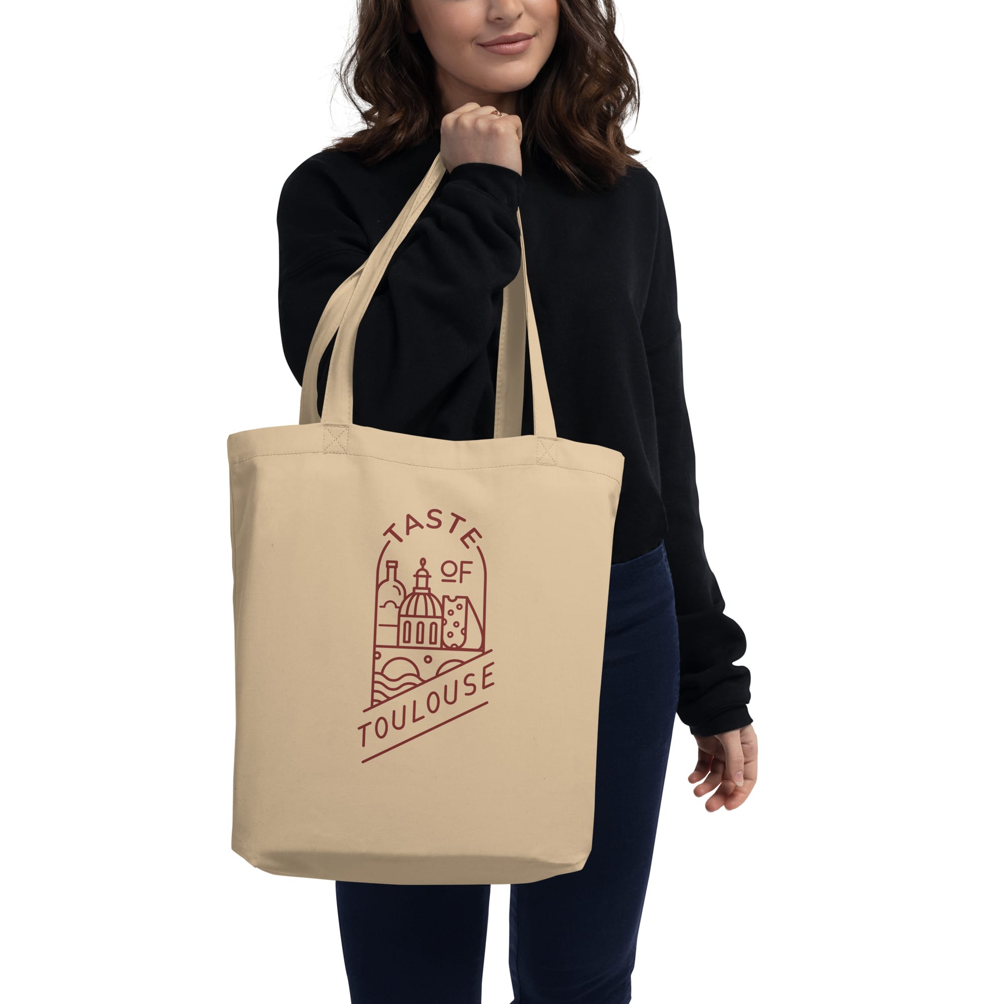 A woman in a black sweater holds the Tote Bag with Red Logo, featuring a red line drawing of Toulouse landmarks and "Taste of Toulouse" printed on beige fabric.