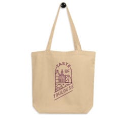 Beige Tote Bag with Wine Logo hangs on a hook, showcasing purple line art of Toulouse landmarks and the phrase "Taste of Toulouse.