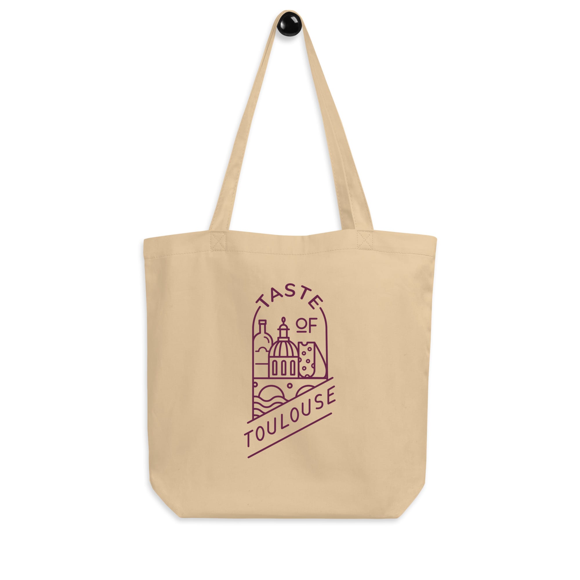 Beige Tote Bag with Wine Logo hangs on a hook, showcasing purple line art of Toulouse landmarks and the phrase "Taste of Toulouse.