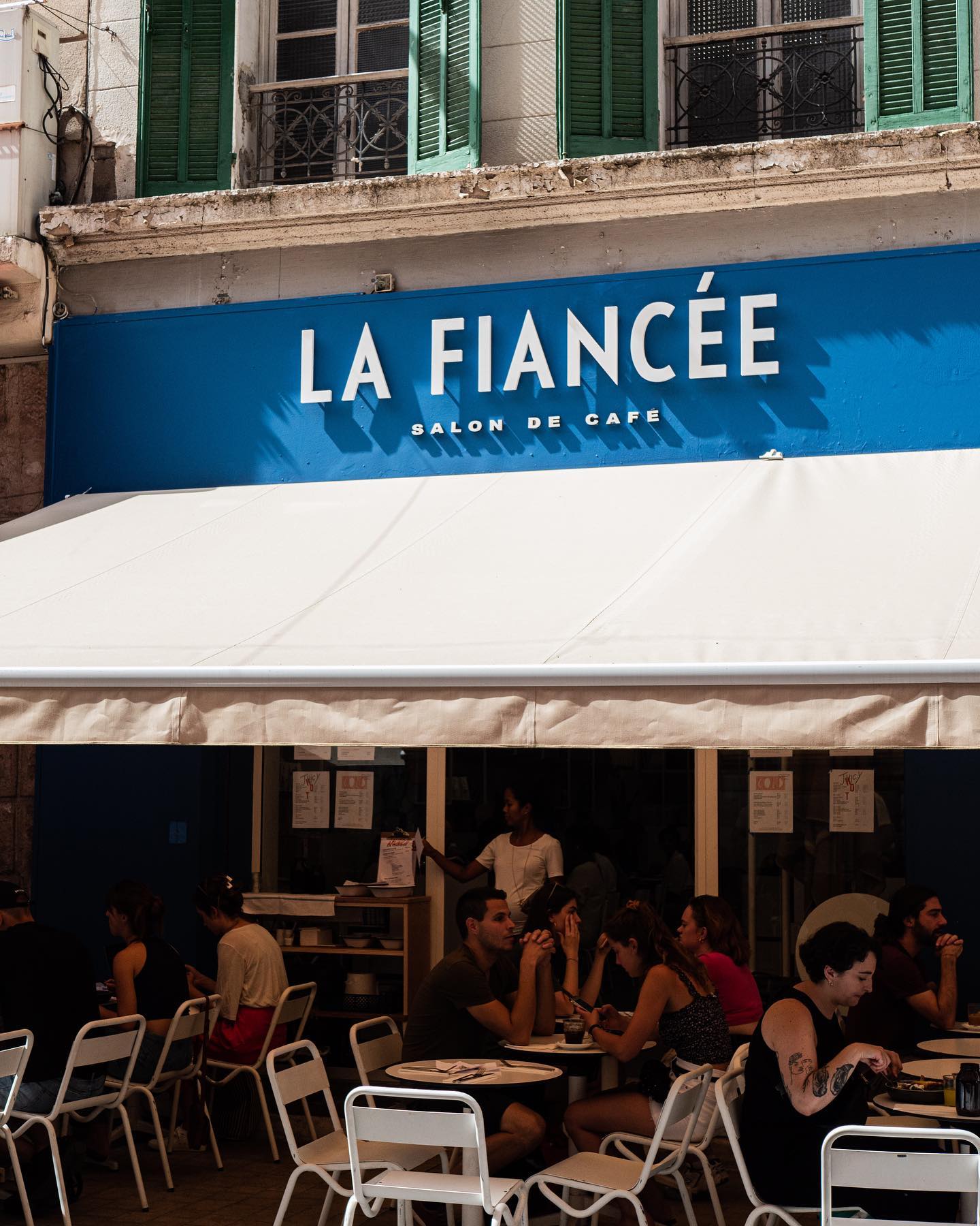 Outdoor seating at "La Fiancée," one of the best coffee shops in Toulouse, features white chairs and tables under a canopy. Guests chat while staff work inside the blue café with white lettering.