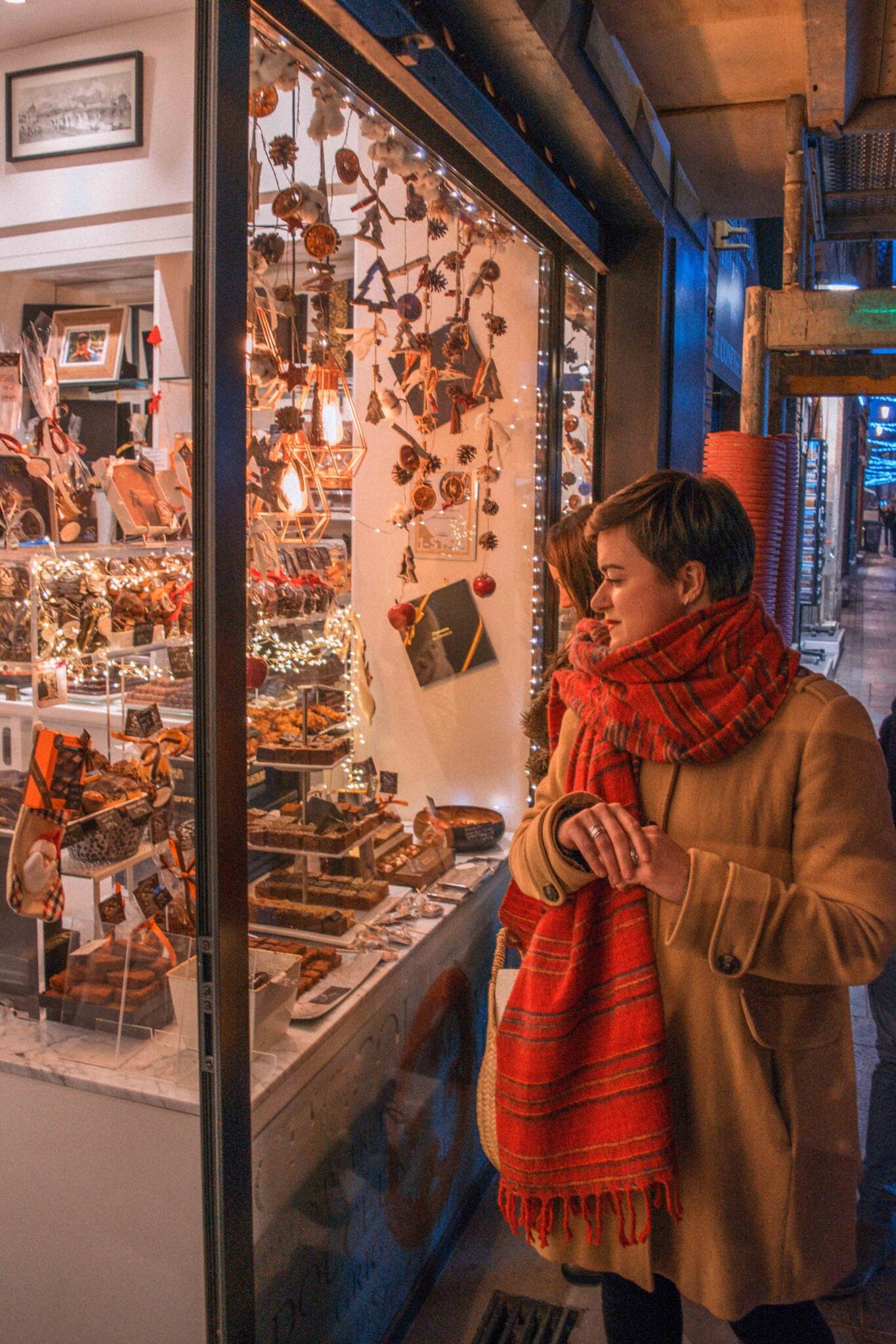 A person in a tan coat and red scarf stands outside a festive shop window in Toulouse, gazing at the Christmas lights, ornaments, and treats inside—a perfect addition to your list of things to do this holiday season.