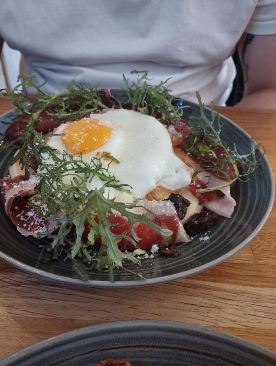 A person in a white shirt sits at a table with a plate of food topped with a sunny-side-up egg, cured meat, greens, and grated cheese. Served in a dark bowl, it's the perfect start to a Toulouse Brunch experience.