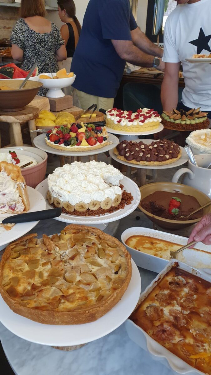 A table crowded with various desserts and baked dishes, including pies, cakes, and tarts, captures the spirit of the best brunches in Toulouse as people in casual clothing serve themselves in the background.