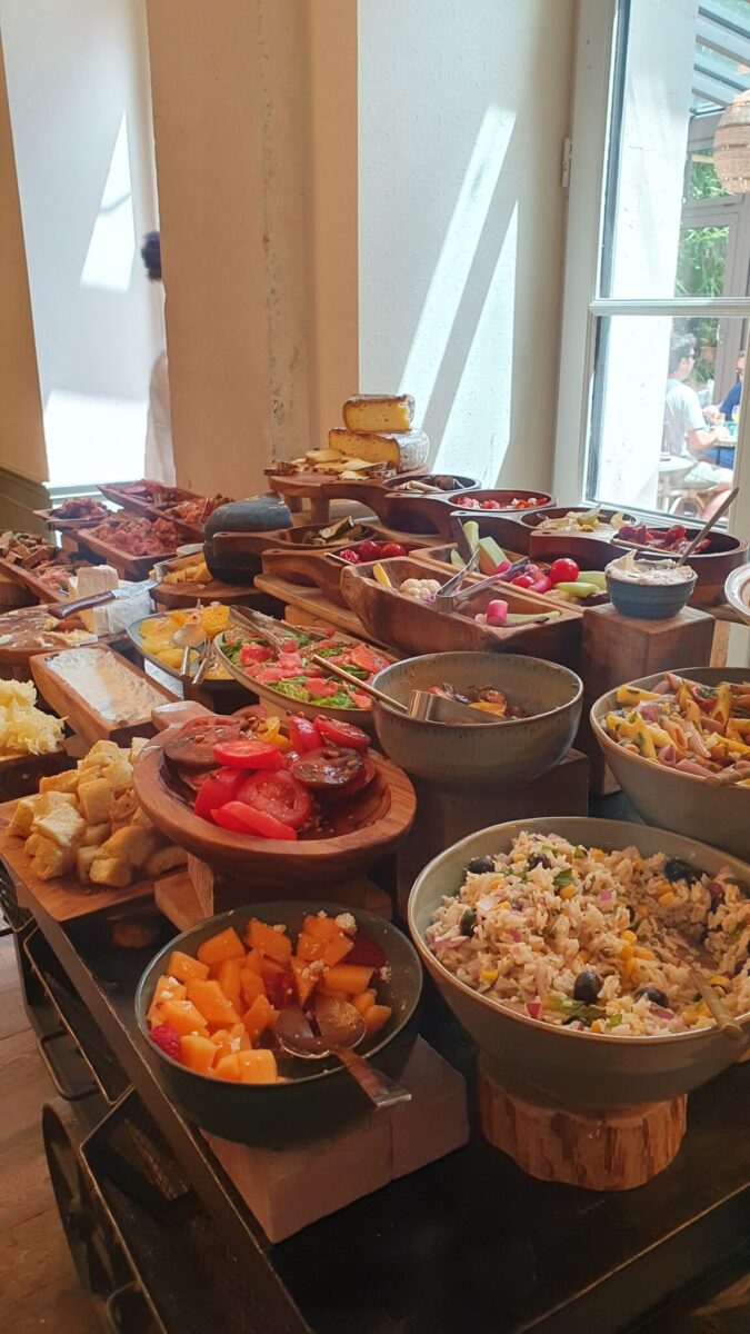 A buffet table filled with various dishes—salads, fresh fruit, bread, cheese, sliced meats, and cakes—all arranged in bowls and wooden trays near a sunlit window, captures the inviting spirit of the best brunches in Toulouse.