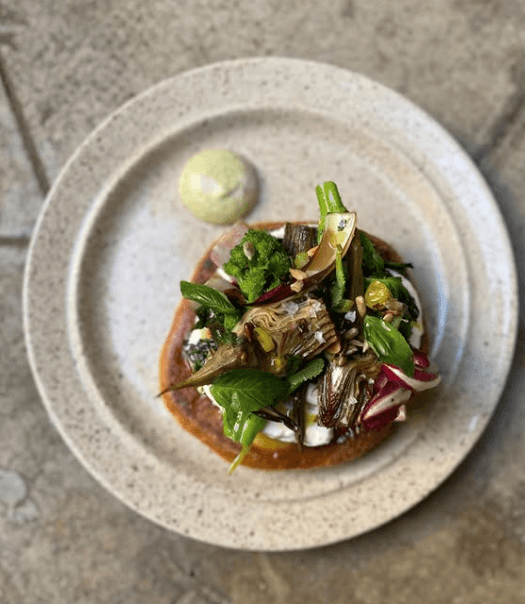 A round plate holds a flatbread topped with fresh greens, vegetables, and herbs, with a small dollop of light-colored sauce on the side—a fresh option often found at the best brunches in Toulouse, set on a stone or tile surface.