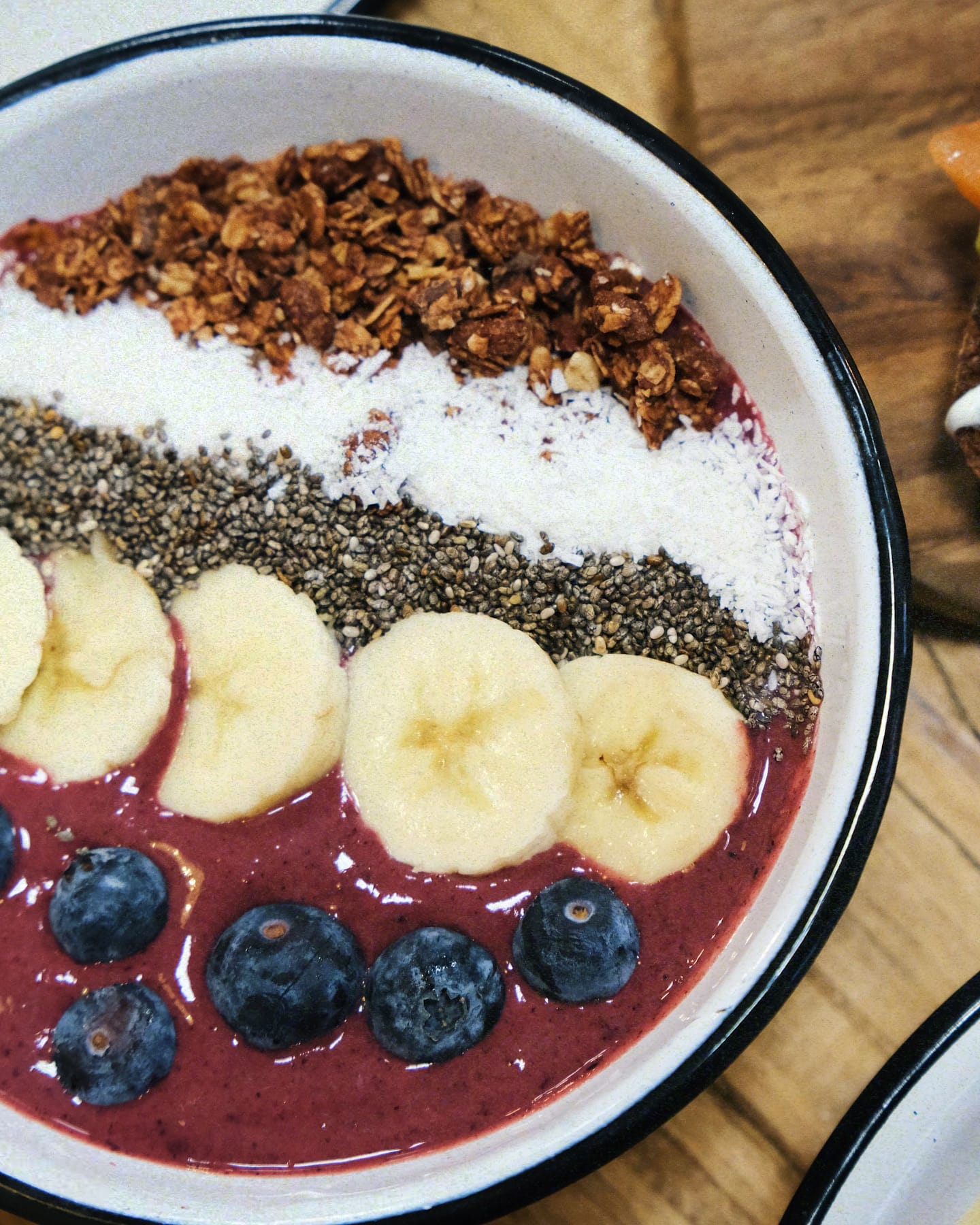 A smoothie bowl topped with rows of granola, shredded coconut, chia seeds, banana slices, and blueberries, served in a white bowl with a black rim on a wooden surface—perfect for those seeking the best brunches in Toulouse.