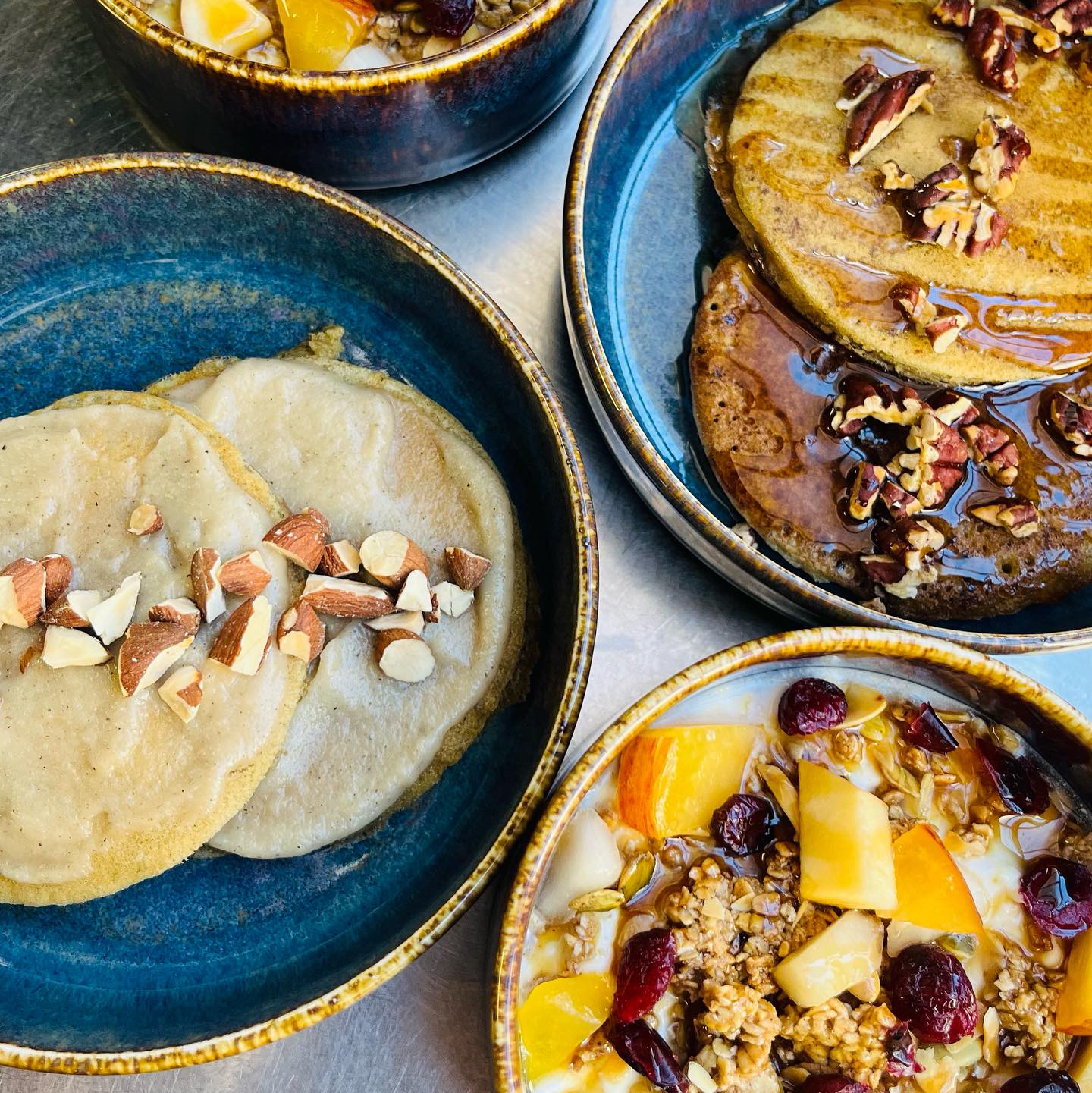 Four ceramic bowls hold delicious breakfast foods: pancakes with chopped nuts and syrup, plus granola with fresh fruit and cranberries—like a taste of the best brunches Toulouse has to offer.