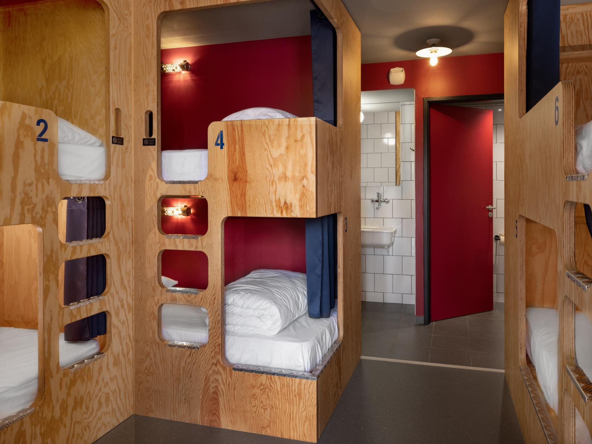 A modern hostel room with wooden bunk beds, white bedding, and blue numbers. Red walls and a door lead to a white-tiled bathroom. Clean, minimalist decor—an excellent choice for Toulouse accommodation.