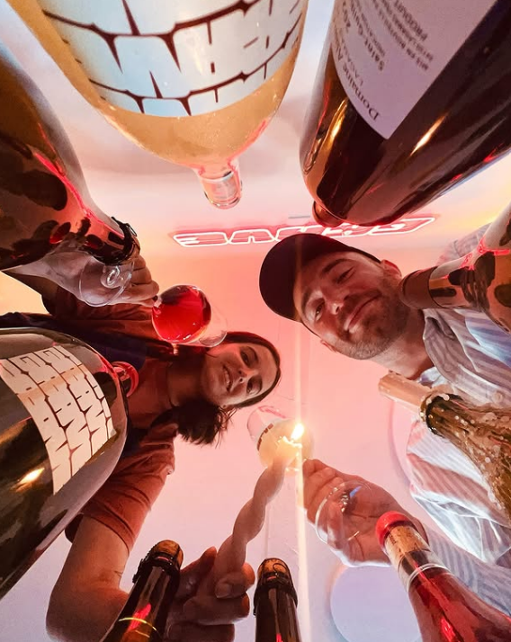 Grave wine bar, Toulouse. Two people seen from below, holding wine glasses and surrounded by several wine bottles in warm lighting at a lively wine bar in Toulouse, smiling and celebrating together beneath a neon sign.