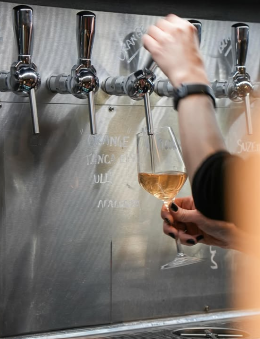 Origin, wine bar. At a lively Toulouse wine bar, a person fills a glass from a metal tap on a wall lined with multiple taps. Wearing a black sleeve and watch, they stand before handwritten notes decorating the metal backdrop.