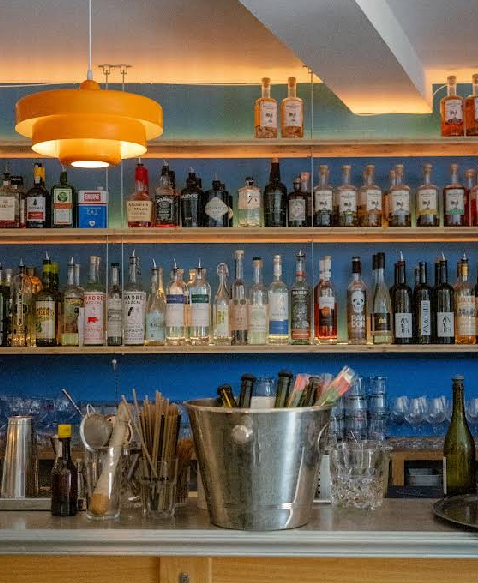 Bistrot 12, wine bar. A bar counter with a metal ice bucket, glasses, and cocktail supplies in the foreground evokes the atmosphere of Toulouse wine bars. Shelves behind display various liquor bottles, illuminated by a modern orange ceiling lamp above.