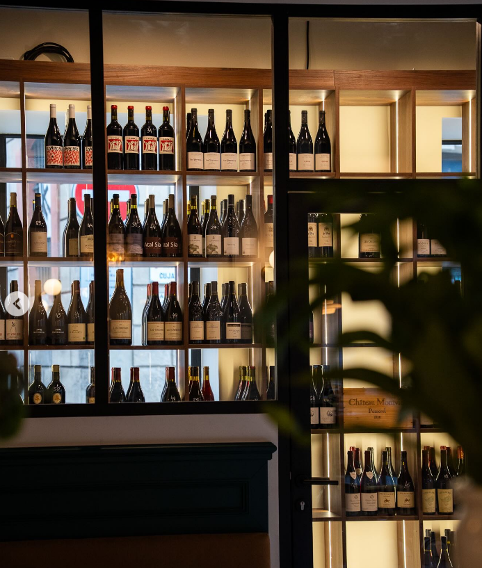 Cafe de la Bourse, wine bar. A well-lit wine cellar/cave filled with various bottles on wooden shelves, seen through glass doors in a cozy, modern interior—evoking the stylish ambiance of wine bars Toulouse is known for. Some greenery is slightly visible in the foreground.