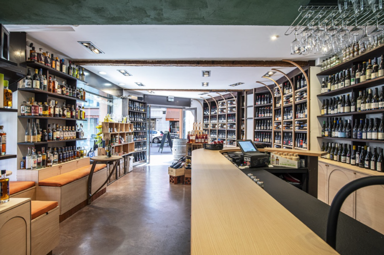 A modern liquor store interior in Toulouse with wooden shelves lined with bottles of wine and spirits, a long counter like those in wine bars, and glassware hanging above. Orange-cushioned benches run along one wall.
