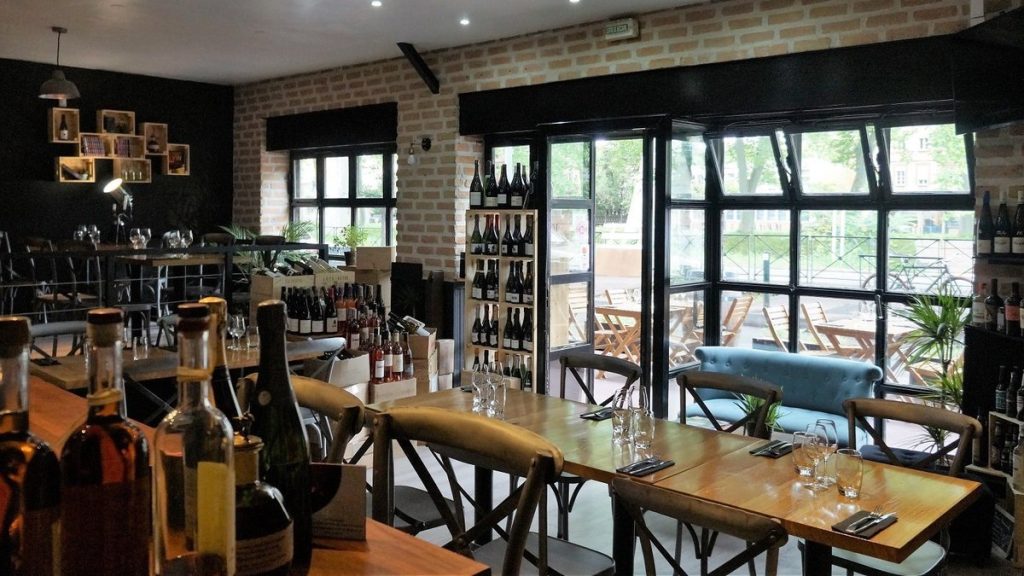 A cozy restaurant interior in Toulouse with wooden tables, chairs, bottles of wine on shelves, and large windows looking out onto an outdoor seating area with greenery—one of the best wine bars in the city.
