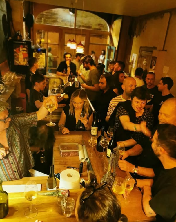 Nabu, Toulouse, wine. A lively bar scene at one of the best wine bars in Toulouse, with people socializing, holding drinks, and smiling around a wooden counter filled with wine glasses and bottles under warm lighting.
