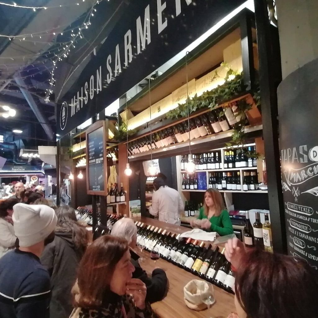 Maison Sarment. A busy wine bar in Toulouse with shelves full of bottles, string lights, and people gathered at the counter enjoying drinks. A bartender in green serves customers under a black sign that reads "HANSON SARMENT.