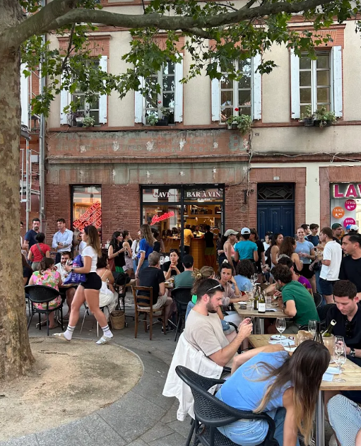 Soulan wine bar and wine shop. A lively outdoor café scene in Toulouse, with people sitting at tables eating and drinking, while others mingle near a leafy tree. The backdrop is an old building with large windows and a stylish wine bar.