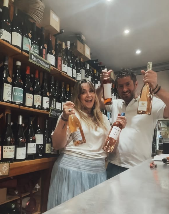 Two people stand behind a counter in a Toulouse wine shop, smiling and holding bottles of rosé wine. Shelves filled with various wines line the wall, giving the space the cheerful, lively vibe found in local wine bars.